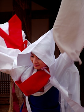 A person is wearing traditional clothing, characterized by flowing white and red garments. The attire appears to be a traditional costume, possibly Korean, with a hooded white top and a vivid red sash. The person shows a gentle smile with poised movement.