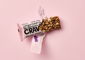 A granola bar partially unwrapped from pink packaging lies on a pink surface. The bar's texture appears nutty, with visible chunks of cashews and other grains.