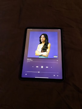 An electronic tablet displaying a music streaming application. The screen shows the cover art of a song, featuring a figure with long hair and a white top against a purple background. The person's face is decorated with colorful stickers. The music player interface is visible with playback controls and track information.