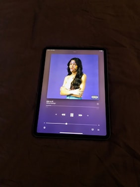 An electronic tablet displaying a music streaming application. The screen shows the cover art of a song, featuring a figure with long hair and a white top against a purple background. The person's face is decorated with colorful stickers. The music player interface is visible with playback controls and track information.