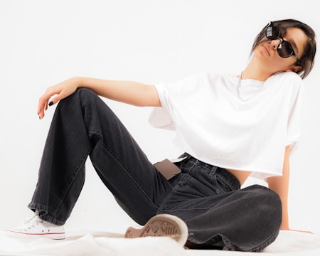 A person is sitting in a relaxed pose wearing oversized black jeans, a loose white t-shirt, and sunglasses. The background is plain white, and the overall pose conveys an air of casual confidence.