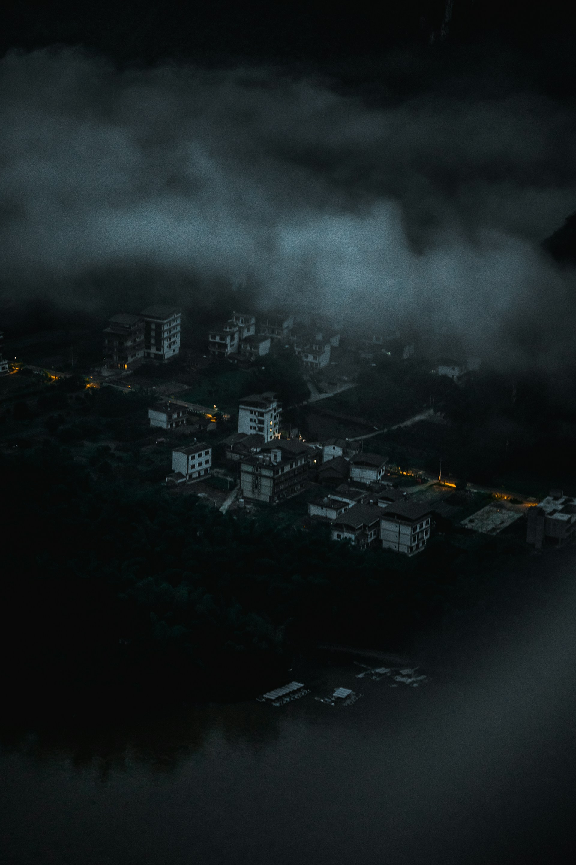 A mist-shrouded village at twilight, with shadowy outlines of traditional nipa huts and twisted trees looming under a heavy fog.