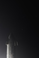 A metallic rocket is standing upright against a dark night sky. It has a sleek, shiny surface with visible seams and panels. The lighting accentuates the reflective properties of its material.