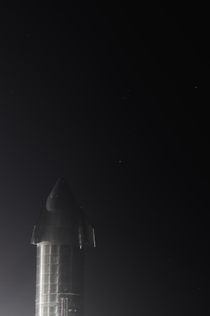 A metallic rocket is standing upright against a dark night sky. It has a sleek, shiny surface with visible seams and panels. The lighting accentuates the reflective properties of its material.