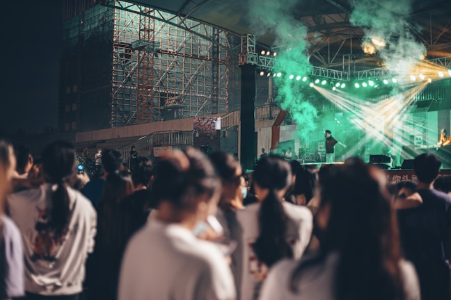 A dimly lit concert scene with a large crowd gathered in front of a stage. The stage is illuminated by bright, green spotlights that create beams of light through the smoke. An artist or performer is on stage, with musical equipment and scaffolding visible in the background.