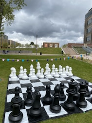 A large outdoor chess set is placed on a checkered mat on a grassy lawn. The chess pieces are oversized, with black pieces in the foreground and white pieces in the background. Surrounding the chess set are areas of grass bordered by modern buildings, stairs, and murals. The sky is overcast with dark, looming clouds, creating a dramatic contrast against the vibrant green grass.
