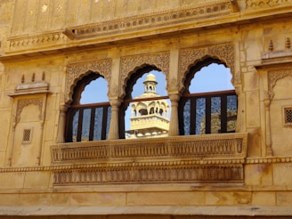 An ornate, carved stone facade features three arched windows, with two reflecting the image of a distant tower against a clear blue sky. The architecture is detailed with intricate patterns and motifs, characteristic of traditional design.