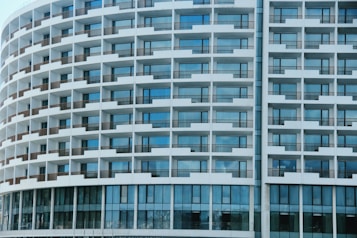 The image features a modern, curved building facade comprising multiple balconies organized in a repeating grid pattern. Each balcony has a railing, and the exterior is predominantly white with blue glass windows. The windows reflect the sky, creating a harmonious blend with the building's clean architectural lines.