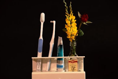 A neatly organized setup includes a blue-and-white toothbrush and an interdental brush in a holder. Beside them, a tube of toothpaste is placed, showcasing a modern and clean aesthetic. To the right, a small glass container holds vibrant yellow flowers and a single red rose, adding a touch of natural beauty.