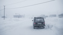 A vehicle is traveling through a snow-covered road during a heavy snowstorm. The visibility is low, and snow heavily blankets the landscape around, with faint outlines of buildings and utility poles visible in the background.