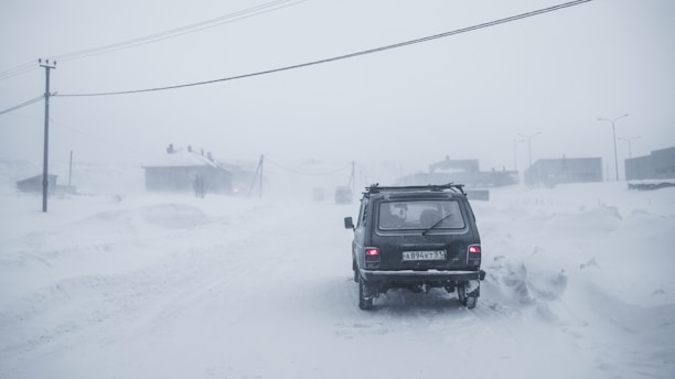 A vehicle is traveling through a snow-covered road during a heavy snowstorm. The visibility is low, and snow heavily blankets the landscape around, with faint outlines of buildings and utility poles visible in the background.