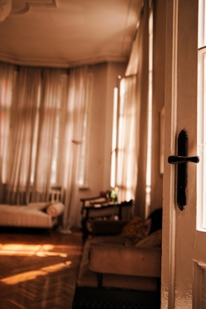 A cozy and sunlit room with large curtained windows allowing warm light to filter in. The room features a wooden parquet floor, a beige sofa with patterned pillows, and a single bed with a neatly placed pillow. Near the center, a small green plant sits on a table. The open door frames the view, adding depth to the space.