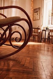 A room featuring a wooden rocking chair with intricate swirls is shown in the foreground. The warm, golden light from the window illuminates the wooden floorboards, and cozy furniture like chairs and a small table appear in the background. A potted plant sits by the window, and a piece of art hangs on the wall.