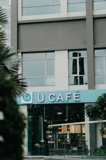 The storefront of a café with a blue sign that reads 'U CAFÉ'. The building features large windows above the entrance and there are some plants visible on the sides. The interior of the café is slightly visible through the glass doors, showing some seating and illuminated menu boards.