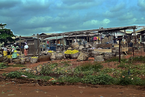 An outdoor market scene with several large woven baskets filled with yellow fruits. People are standing and walking around the market area, which has makeshift wooden structures and stalls. The ground is reddish-brown dirt with some vegetation. The sky above is cloudy.