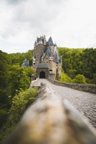 A medieval castle sits atop a hill surrounded by lush green forests. The castle features multiple towers with pointed roofs and intricate details. A cobblestone path leads up to the castle, creating a sense of depth in the image.