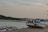 A damaged boat rests on a concrete platform by the shore, showing signs of rust and decay. Nearby, several smaller boats are docked in the calm waters. The background features a lush green landscape and a distant view of a coastal town under a cloudy sky. A bird is visible flying above the water.