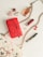 A collection of fashion accessories is artfully arranged on a light surface. Items include a vibrant red handbag with intricate floral embroidery, a white quilted purse with black and pink rose patterns, a gold chain, a red lipstick, a mascara, a ring, small polished stones, and a bottle of perfume. The color palette is dominated by red, pink, gold, and white hues, exuding a sense of elegance and luxury.