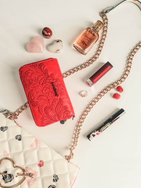 A collection of fashion accessories is artfully arranged on a light surface. Items include a vibrant red handbag with intricate floral embroidery, a white quilted purse with black and pink rose patterns, a gold chain, a red lipstick, a mascara, a ring, small polished stones, and a bottle of perfume. The color palette is dominated by red, pink, gold, and white hues, exuding a sense of elegance and luxury.