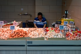 A man is seated behind a stall displaying various types of eggs in cardboard trays. The eggs are sorted by color and size, with several price tags visible. The stall is indoors, with a tiled wall and electronic equipment around.