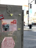 A large gray metal box on the street with several taped notices and stickers attached. One notice warns of potential danger from 'protestors with guns' and advises avoiding certain areas, while another sticker poses a question about actions during the rise of fascism. The blurred background shows an urban environment with traffic lights and indistinct buildings.