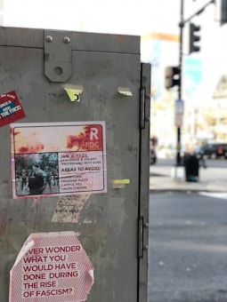 A large gray metal box on the street with several taped notices and stickers attached. One notice warns of potential danger from 'protestors with guns' and advises avoiding certain areas, while another sticker poses a question about actions during the rise of fascism. The blurred background shows an urban environment with traffic lights and indistinct buildings.