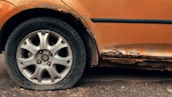 A close-up view of a car wheel with a flat tire, surrounded by a rusted and deteriorating car body. The vehicle's orange paint is peeling and the exposed areas are heavily corroded.