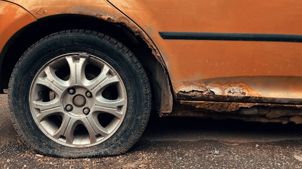 A close-up view of a car wheel with a flat tire, surrounded by a rusted and deteriorating car body. The vehicle's orange paint is peeling and the exposed areas are heavily corroded.