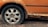 A close-up view of a car wheel with a flat tire, surrounded by a rusted and deteriorating car body. The vehicle's orange paint is peeling and the exposed areas are heavily corroded.