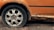 A close-up view of a car wheel with a flat tire, surrounded by a rusted and deteriorating car body. The vehicle's orange paint is peeling and the exposed areas are heavily corroded.