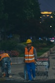 Two construction workers are wearing high-visibility safety vests and helmets while working outdoors at night. One is standing next to a machine, possibly a generator, while the other is bending over to pick up or adjust something. The background is a park-like setting with trees and flowers, and some building lights are visible in the distance.