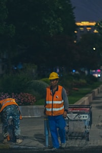 Two construction workers are wearing high-visibility safety vests and helmets while working outdoors at night. One is standing next to a machine, possibly a generator, while the other is bending over to pick up or adjust something. The background is a park-like setting with trees and flowers, and some building lights are visible in the distance.