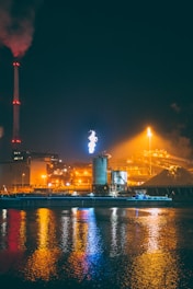 An industrial facility is illuminated against the night sky, with structures including chimneys, towers, and buildings. Bright lights and reflections create vibrant colors on the water surface in the foreground. Smoke can be seen coming from a tall stack, and a flare is burning in the distance.