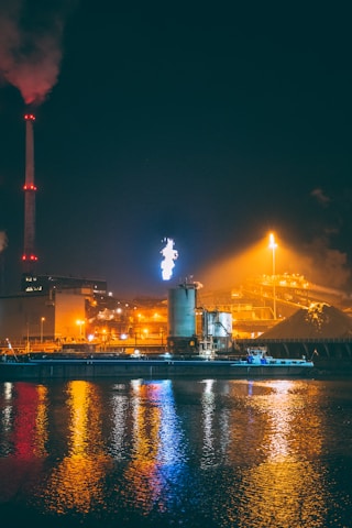 An industrial facility is illuminated against the night sky, with structures including chimneys, towers, and buildings. Bright lights and reflections create vibrant colors on the water surface in the foreground. Smoke can be seen coming from a tall stack, and a flare is burning in the distance.