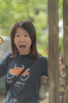 A person with an expression of surprise or excitement is captured in a casual outdoor setting. The individual has long dark hair and is wearing a blue t-shirt with a bicycle graphic and the words 'Live Simply'. The background features soft focus greenery suggesting a park or natural area.