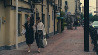 Two women walk along a quiet, urban street lined with buildings. One woman, in dark clothing, carries a shopping bag, while the other, dressed in a light blouse and patterned skirt, looks at her phone. The street appears to be calm, with a few other pedestrians in the distance.