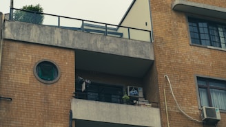 The facade of a multi-story building with a balcony and several windows. The building has a plain stone exterior with a circular window and a balcony featuring potted plants and an air conditioning unit. An iron railing and some clothes hanging to dry are visible.