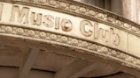 An old, weathered sign above an entrance labeled 'Music Club' is adorned with intricate floral carvings. The sign has a vintage appearance, with chipped and faded letters and a surface showing signs of aging.