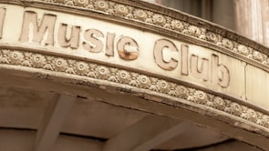 An old, weathered sign above an entrance labeled 'Music Club' is adorned with intricate floral carvings. The sign has a vintage appearance, with chipped and faded letters and a surface showing signs of aging.