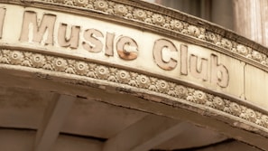 An old, weathered sign above an entrance labeled 'Music Club' is adorned with intricate floral carvings. The sign has a vintage appearance, with chipped and faded letters and a surface showing signs of aging.