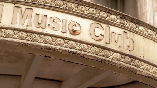 An old, weathered sign above an entrance labeled 'Music Club' is adorned with intricate floral carvings. The sign has a vintage appearance, with chipped and faded letters and a surface showing signs of aging.