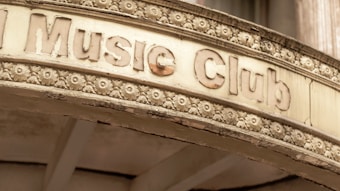 An old, weathered sign above an entrance labeled 'Music Club' is adorned with intricate floral carvings. The sign has a vintage appearance, with chipped and faded letters and a surface showing signs of aging.