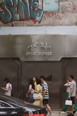 A group of people are walking past a modern cafe with a metallic facade labeled 'Cyber Coffee'. The background features graffiti art and a partially visible SPA sign. There is also a parked black car in the foreground.