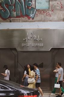 A group of people are walking past a modern cafe with a metallic facade labeled 'Cyber Coffee'. The background features graffiti art and a partially visible SPA sign. There is also a parked black car in the foreground.