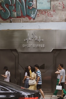 A group of people are walking past a modern cafe with a metallic facade labeled 'Cyber Coffee'. The background features graffiti art and a partially visible SPA sign. There is also a parked black car in the foreground.