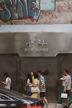 A group of people are walking past a modern cafe with a metallic facade labeled 'Cyber Coffee'. The background features graffiti art and a partially visible SPA sign. There is also a parked black car in the foreground.