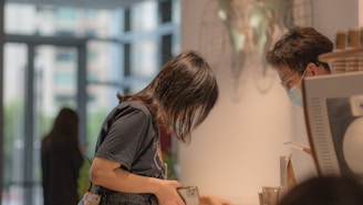 A woman is interacting with a man at a counter. The woman is holding a smartphone and appears to be engaging in some form of communication or transaction. The man is wearing a face mask, which indicates awareness of health and safety protocols. In the background, there is a blurred figure and some large windows through which outdoor scenery is visible.