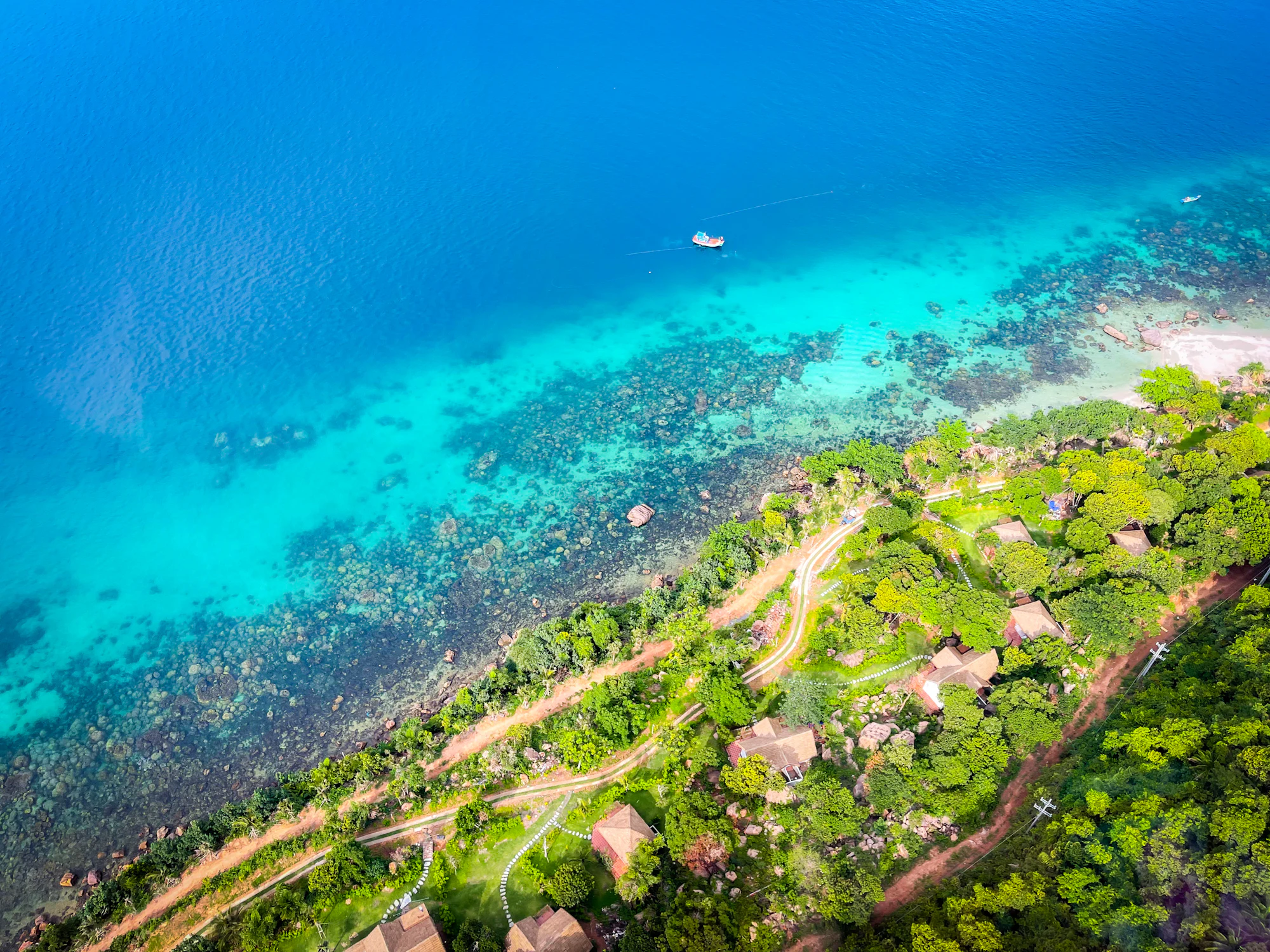 The Phu Quoc coast from the air — pale turquoise shallows over reef, a wooded green shoreline, and a single boat out in the deeper blue