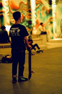 A person holding a skateboard is standing in an indoor area with vibrant, psychedelic wall art in the background. Other people are sitting and engaging with each other in the same area.