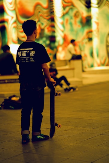 A person holding a skateboard is standing in an indoor area with vibrant, psychedelic wall art in the background. Other people are sitting and engaging with each other in the same area.
